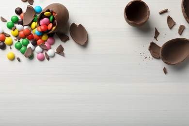 Broken chocolate eggs and colorful candies on white wooden table, flat lay. Space for text