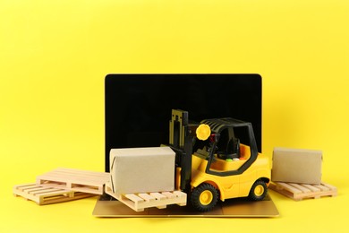 Laptop, toy forklift with wooden pallets and boxes on yellow background