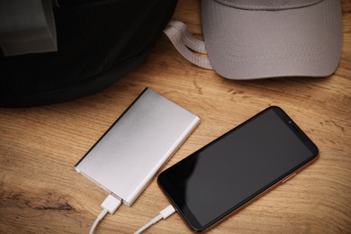Photo of Smartphone charging with power bank, backpack and hat on wooden table
