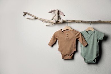Baby bodysuits and toy on decorative branch near light wall. Space for text