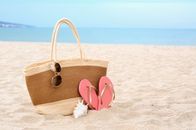 Stylish straw bag with sunglasses, flip flops and shell on sand near sea, space for text. Beach accessories