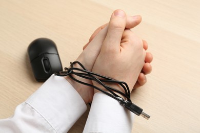 Photo of Man showing hands tied with computer mouse cable at wooden table, closeup. Internet addiction