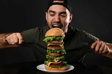 Photo of Young hungry man with cutlery eating huge burger on black background