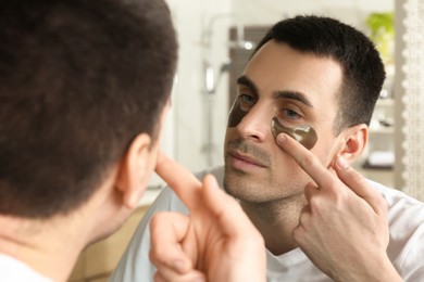 Young man applying under eye patches near mirror at home