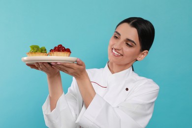 Photo of Happy professional confectioner in uniform with delicious tartlets on light blue background