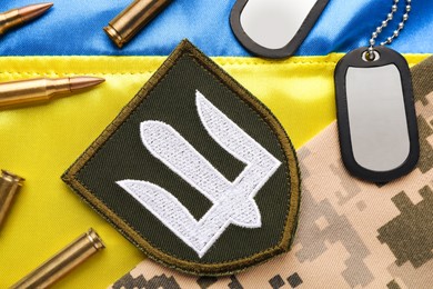 Ukrainian army chevron near camouflage fabric, bullets and military ID tags on national flag of Ukraine, flat lay