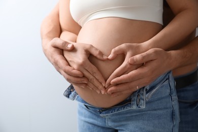 Pregnant young woman making heart with hands on belly and husband near her against light background, closeup
