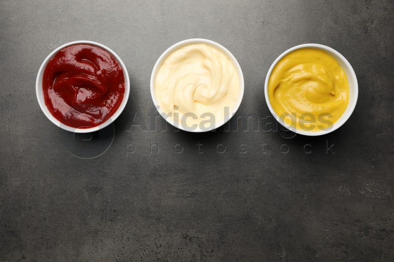 Ketchup, mustard and mayonnaise in bowls on grey table, flat lay. Space for text