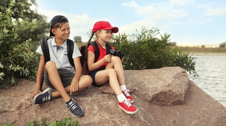 Cute little children with backpacks sitting on rock near river. Camping trip