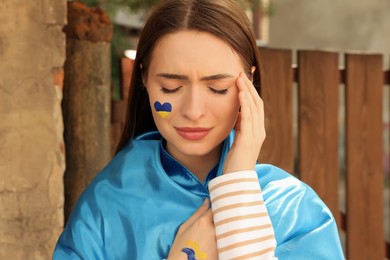 Photo of Sad young woman with drawing of Ukrainian flag on face outdoors