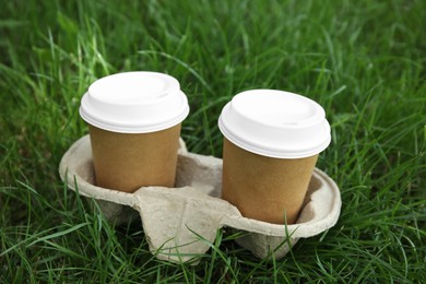 Takeaway paper coffee cups with plastic lids in cardboard holder on green grass outdoors
