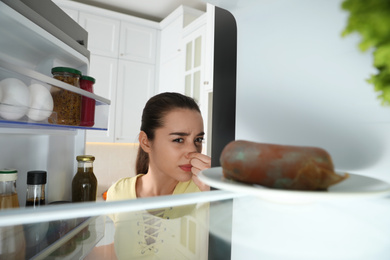 Young woman feeling bad smell of spoiled sausage in refrigerator, view from inside