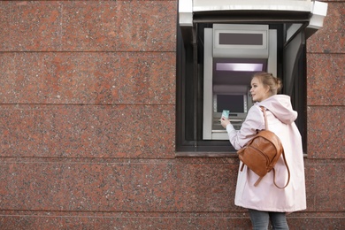 Young woman with credit card near cash machine outdoors. Space for text