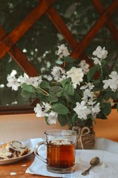 Glass cup of aromatic tea, tasty dessert and beautiful jasmine flowers on wooden table indoors