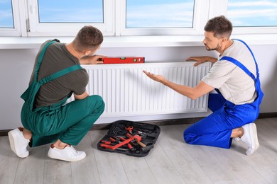 Professional plumbers using bubble level for installing new heating radiator in room