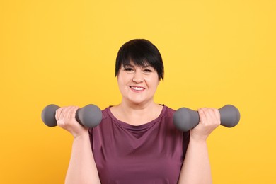 Happy overweight mature woman doing exercise with dumbbells on yellow background