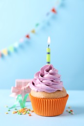 Birthday cupcake with burning candle and gift box on light blue table