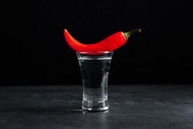 Photo of Red hot chili pepper and vodka in shot glass on grey table against black background