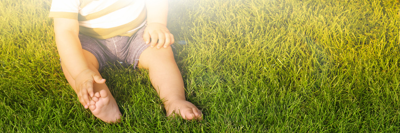 Adorable little baby sitting on green grass outdoors, space for text. Banner design