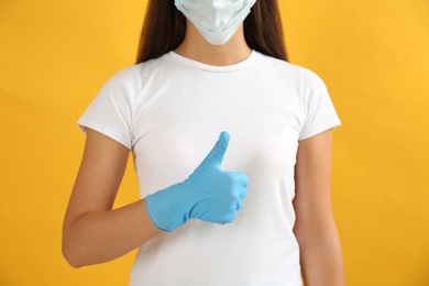 Woman in protective face mask and medical gloves showing thumb up gesture on yellow background, closeup