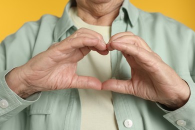 Elderly woman making heart with her hands on yellow background, closeup