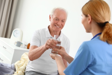 Nurse giving glass of water to elderly man indoors. Medical assistance