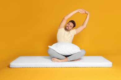 Man with pillow sitting on soft mattress and stretching against orange background