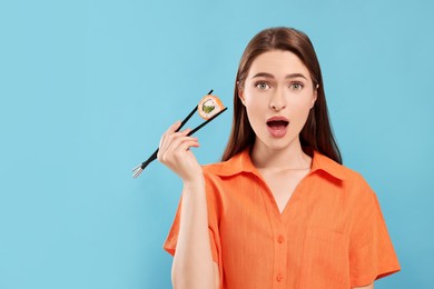 Photo of Emotional young woman holding sushi roll with chopsticks on light blue background. Space for text