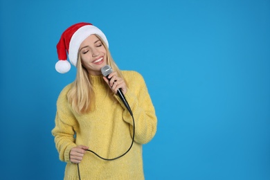 Happy woman in Santa Claus hat singing with microphone on blue background, space for text. Christmas music