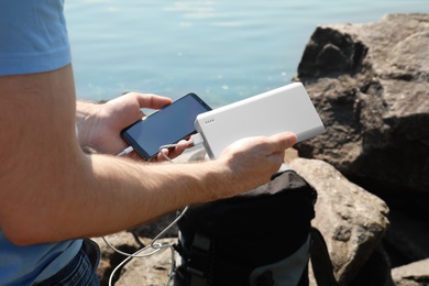 Photo of Man charging mobile phone with power bank on rocky mountain near river, closeup