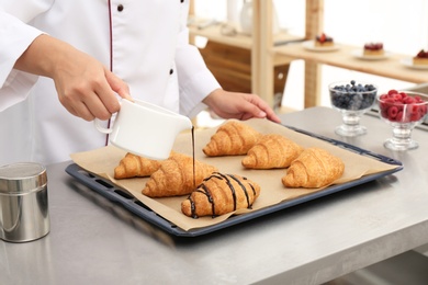 Photo of Young female pastry chef pouring chocolate sauce onto croissants at table in kitchen, closeup