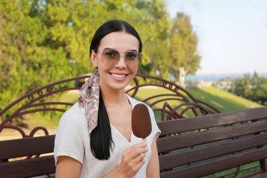 Beautiful young woman holding ice cream glazed in chocolate on bench outdoors
