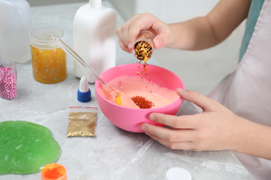 Photo of Little girl adding colored sparkles into homemade slime toy at table, closeup of hands
