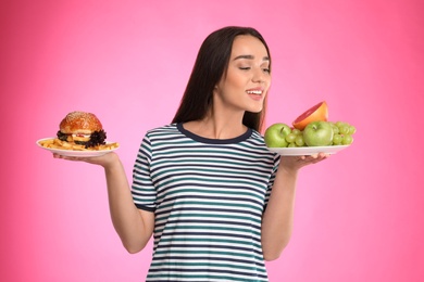 Woman choosing between fruits and burger with French fries on pink background