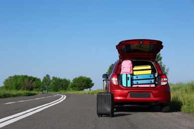 Suitcase near family car with open trunk full of luggage on highway. Space for text
