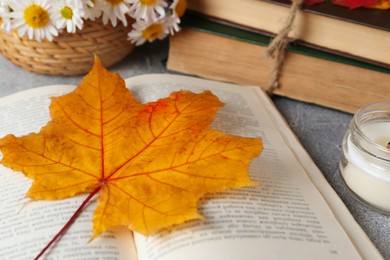 Photo of Book with autumn leaf as bookmark and scented candle on light gray table, closeup