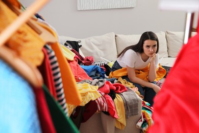 Photo of Upset young woman with lots of clothes on sofa in room. Fast fashion