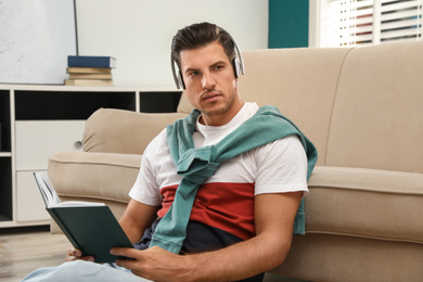 Man with headphones and book near sofa indoors. Audiobook concept
