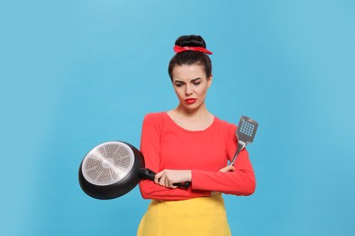 Housewife with frying pan and spatula on light blue background
