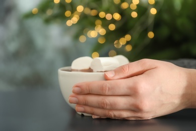Woman holding cup of aromatic cacao with marshmallows on table, closeup. Space for text