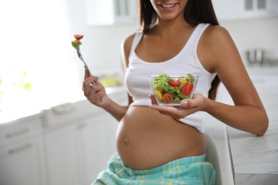 Young pregnant woman with bowl of vegetable salad at table in kitchen, closeup. Taking care of baby health