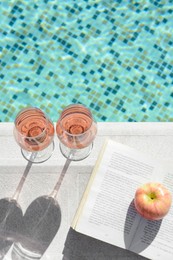 Photo of Glasses of tasty rose wine, open book and apple on swimming pool edge
