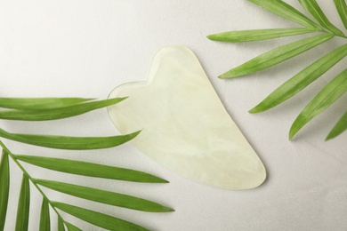 Photo of Jade gua sha tool and green leaves on grey table, flat lay