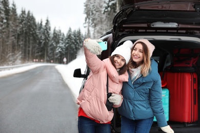 Friends taking selfie near open car trunk full of luggage on road, space for text. Winter vacation