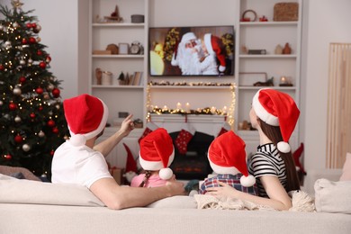 Photo of Family wearing Christmas hats watching TV in cosy room, back view. Winter holidays atmosphere