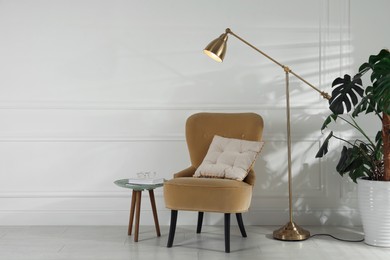 Photo of Comfortable armchair, table, floor lamp and houseplant near white wall in room, space for text. Interior design