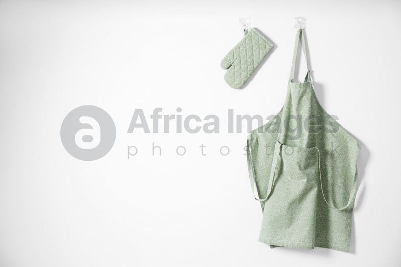 Photo of Clean apron with pattern, kitchen tools and oven glove on light wall. Space for text