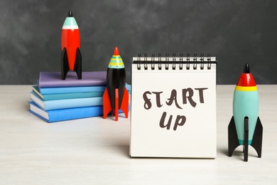 Photo of Notebook with phrase Start Up, toy rockets and notebooks on white table