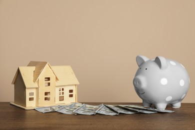 Photo of Piggy bank, dollar banknotes and house model on wooden table against beige background