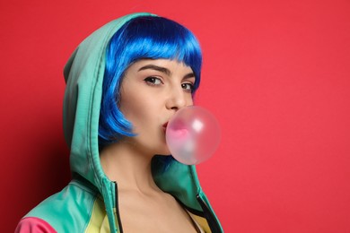 Fashionable young woman in colorful wig blowing bubblegum on red background, space for text